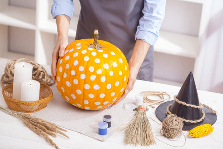 Woman in apron painting Halloween pumpkin, at wooden table, preparing holiday decorations at homeの写真素材