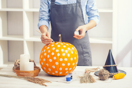 Woman in apron painting Halloween pumpkin, at wooden table, preparing holiday decorations at homeの写真素材
