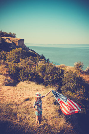 Beautiful young girl holding an American flag on the wind in a meadow. Summer landscape against the blue sky. Independence Day Fourth of Julyの写真素材
