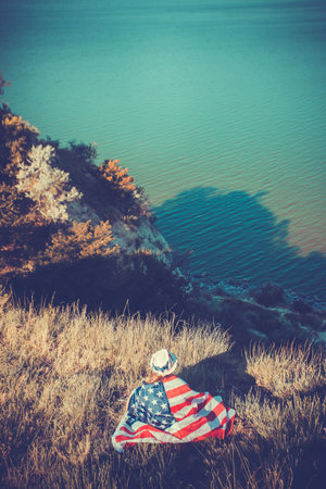 Beautiful young girl holding an American flag on the wind in a meadow. Summer landscape against the blue sky. Independence Day Fourth of Julyの写真素材