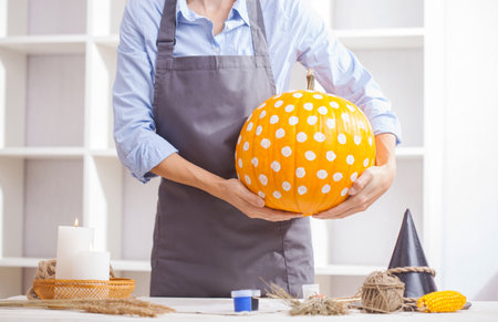 Woman in apron painting Halloween pumpkin, at wooden table, preparing holiday decorations at homeの写真素材