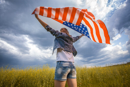 Beautiful young girl holding an American flag on the wind in a rapeseed field. Summer landscape against the blue sky. Independence Day Fourth of Julyの写真素材