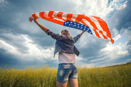 Beautiful young girl holding an American flag on the wind in a rapeseed field. Summer landscape against the blue sky. Independence Day Fourth of Julyの写真素材
