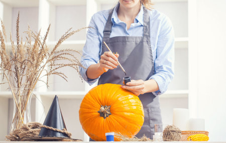 Woman in apron painting Halloween pumpkin, at wooden table, preparing holiday decorations at homeの写真素材
