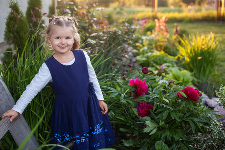 cheerful 3-year-old girl in a blue dress walks near the flowering flower bed in the backyard. happy...