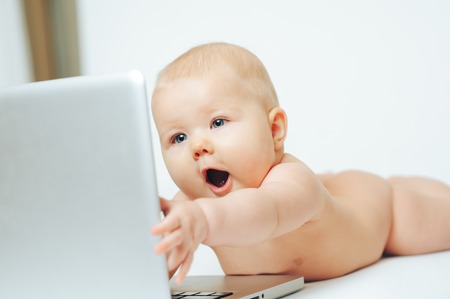 A baby with a surprised face lies in front of a silver laptop and holds out his handの写真素材
