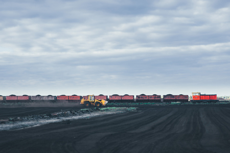 Yellow tractor rides on the field and picks up a black dust. rural the celestial horizon. tractor ahead of the train. train carrying cars filled with sand and soil.の写真素材