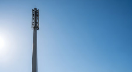 A tall cell tower stands against a vast, clear blue sky, with a bright sun flare visible on the left side.の素材