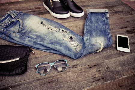 Fashionable ripped jeans, sunglasses, bag and footwear on a wooden background.の写真素材