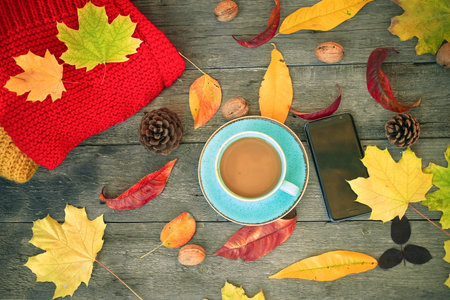 Cup with coffee, knitted warm clothes, autumn leaves and the smartphone on an old wooden background....