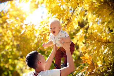 Happy father throws his adorable baby up in autumn park. Father and son spend a day together.の写真素材
