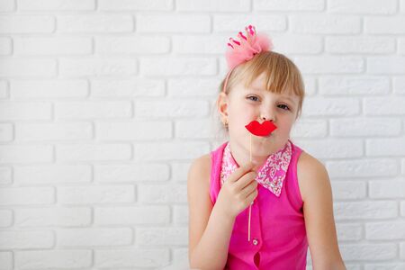 Pretty little girl,  wearing on pink dress, posing with red paper lips, with a crown on his head on white background, waist up. Little Princessの写真素材