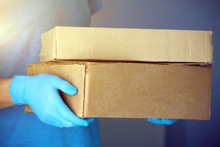 Ð¡ourier man delivers the package to the door, contact less delivery. Male hands in blue rubber gloves hold a cardboard box. Quarantine, safe home deliveryの写真素材