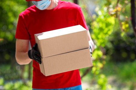 A man in a red T-shirt and black rubber gloves and a mask holds corton boxes in his hands, outdoor....