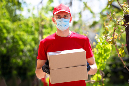 Courier in a red T-shirt and a cap in a medical mask and gloves holds a cardboard box in your hands,...