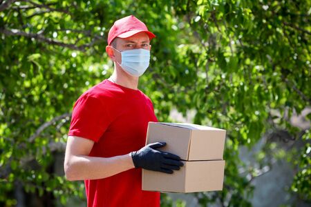 A delivery worker in red uniform holds out cardboard boxes in medical rubber gloves and a mask. Fast and free delivery of transport. Online shopping and express delivery. Quarantineの写真素材