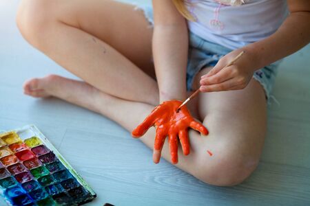 The child sits on the floor and paints his palm with orange paint. Quarantine.  Stay at homeの写真素材