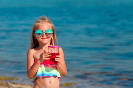 Beautiful little girl in a swimsuit and sunglasses stands by the sea and holds fruit drinks from a glass in her hands. Copy spaceの写真素材