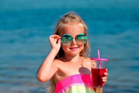 Beautiful little girl in a swimsuit and sunglasses stands by the sea and holds fruit drinks from a glass in her hands. Copy spaceの写真素材