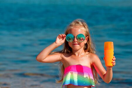 Beautiful little girl in a swimsuit and sunglasses stands by the sea and holds a sunscreen in her hands. Copy spaceの写真素材
