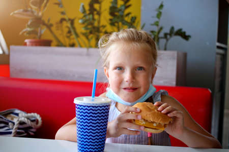 A little girl sits in a cafe in a medical mask and holds a burger and soda in her hands. Opening a cafe. Work cafe with security measures for Covid-19. Work during covid-19 or coronavirusの写真素材