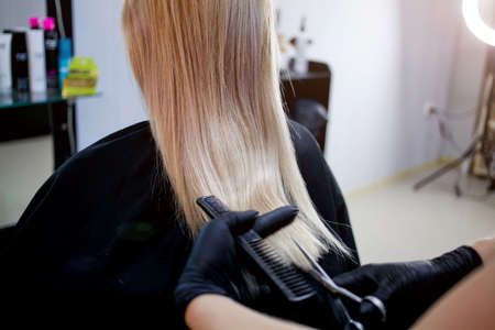 A hairdresser in rubber gloves holds a pair of scissors and a comb. Woman getting a new haircut. Female hairstylist cutting long blond hair with scissors in hair salonの写真素材