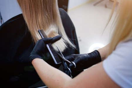 A hairdresser in rubber gloves holds a pair of scissors and a comb. Woman getting a new haircut. Female hairstylist cutting long blond hair with scissors in hair salonの写真素材