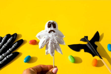 Female hand holding homemade halloween ghost candy on yellow background. Halloween holiday concept