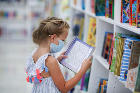 A little girl stands in a bookstore in a medical mask and reads. Child in the library.の写真素材