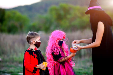 Children trick or treat in Halloween costume and medical mask. A boy and a girl in suits during the coronavirus pandemic receive candy from a woman dressed as a witch.の写真素材