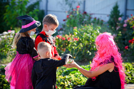 Children trick or treat in Halloween costume and medical mask. A little boy, girl and baby in suits during the coronavirus pandemic receive candy from a woman in a pink wig.の写真素材