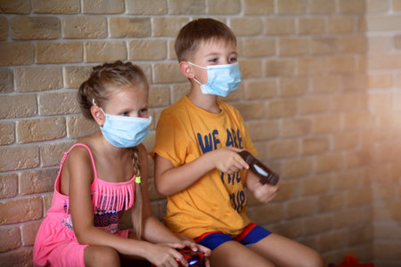 Little girl and boy play medical mask game console while sitting on sofa at home. Corona, covid19,...