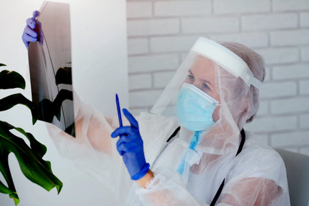 The doctor in PPE looks thoughtfully at the X-ray. Woman doctor in medical mask and protective suit...