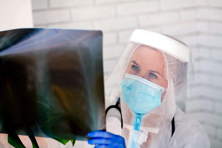 The doctor in PPE looks thoughtfully at the X-ray. Woman doctor in medical mask and protective suit...