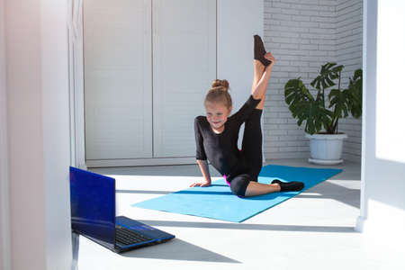 Little girl doing yoga raised her leg up and watching online lessons on laptop while exercising in the room. A child plays sports during COVID-19の写真素材