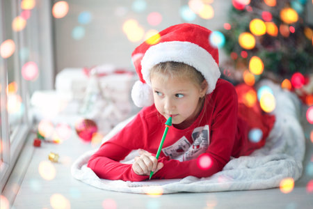 A smiling little girl in a Santa hat writes a letter, dreams of gifts to Santa Claus. Child lying on a blanket on the floor of a house with christmas lightsの写真素材
