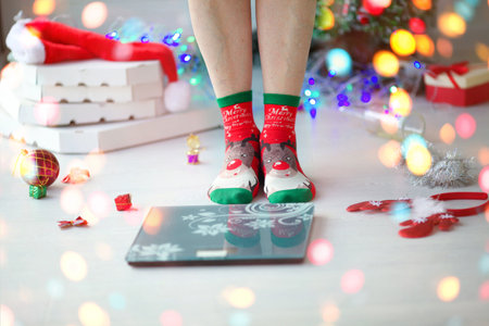 Close-up of adult woman legs in socks with deer stand on scales measure weight. After the holiday Christmas, pizza boxes and New Year's decorations, bottles are lying on the floor. Human measuringの写真素材