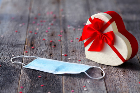 Beautiful holiday box in the shape of a heart with a red ribbon and a medical mask on a wooden background. Valentine's Day during the COVID-19 pandemic. Copy space. The 14th of Februaryの写真素材