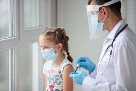 A doctor in a protective suit makes the child in a medical mask a shoulder shot. vaccination.  vaccine. Doctor vaccinating child. Kids at clinic. Little girl getting flu shot.の写真素材
