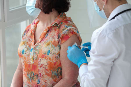 A male doctor in rubber gloves holds a syringe and gives an injection to an older patient wearing a medical mask.の写真素材