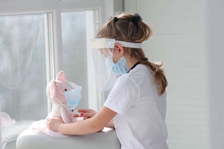 A cute little girl in a medical mask holds a syringe while vaccinating a toy . Smart child playing hospital game pretending to be doctor, nurse concept, veterinarianの写真素材