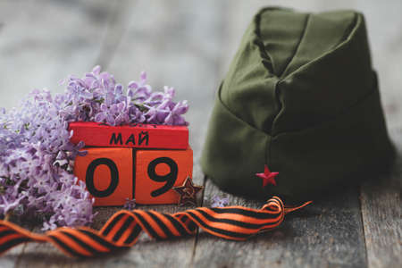 Wooden calendar with Russian text May 9, garrison cap, george ribbon and a bouquet of lilacs on a wooden background. Victory Day.の写真素材