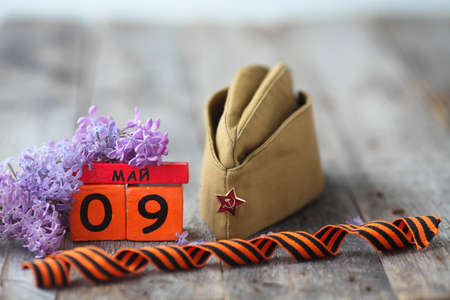Wooden calendar with Russian text May 9, garrison cap, george ribbon and a bouquet of lilacs on a wooden background. Victory Day.の写真素材
