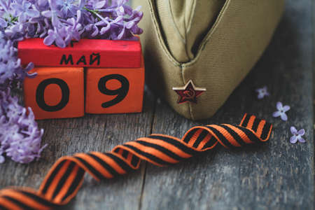 Wooden calendar with Russian text May 9, garrison cap, george ribbon and a bouquet of lilacs on a wooden background. Victory Day.の写真素材
