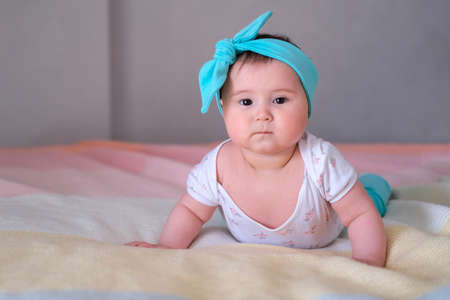 Baby at 6 months raises his body and learns to crawl. A little girl lies on her stomach on the bed and while looking at the camera. A beautiful headband for a child.の写真素材