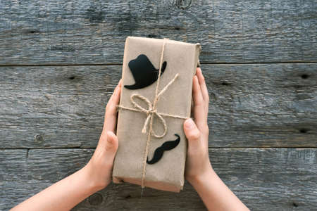 Happy Fathers Day concept. Children is hands hold a gift box for dad made of craft paper on a wooden background. Father's day. Black hat and mustache sign.の写真素材