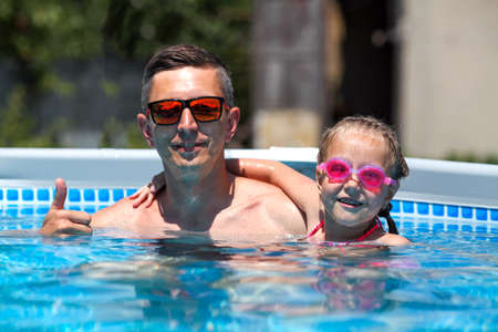 Father in sunglasses swims in the pool with his daughter. Dad and little girl in swimming goggles swim in the pool. A happy man and a child in the water.の写真素材
