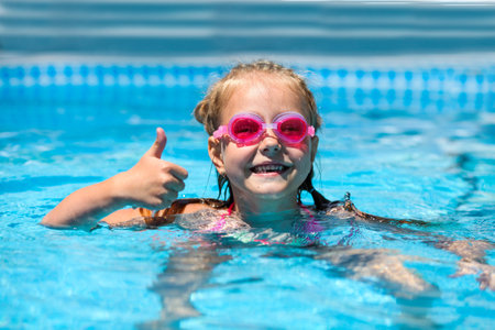 Cute little girl smiling in goggles in the pool on a sunny day. The child showing thumbs up like gesture looking camera. Summer vacationの写真素材