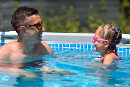Father in sunglasses swims in the pool with his daughter. Dad and little girl in swimming goggles swim in the pool. A happy man and a child in the water.の写真素材