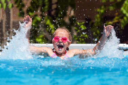 Cute little girl smiling in goggles in the pool on a sunny day. The child splashing water in the pool. Summer vacationの写真素材
